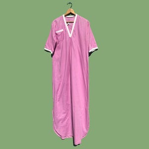 May include: A pink, floor-length nightgown with short sleeves, a V-neck, and a white trim. The nightgown has a pocket on the left side and is hanging on a wooden hanger against a green background.