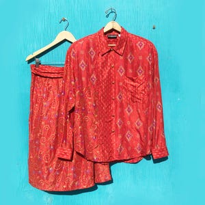 May include: A two-piece red silk outfit, consisting of a long-sleeved button-down shirt and a skirt, hanging on wooden hangers against a turquoise background. The shirt and skirt feature a repeating diamond pattern with floral accents.