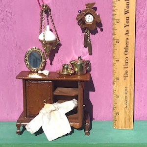May include: A miniature wooden cabinet with a white lace garment hanging on the front, a gold-toned oval mirror, a small teapot, a cup on a saucer, a hanging lamp, and a wall clock. A wooden ruler shows the scale, measuring up to 7 inches in height.