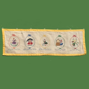 May include: A rectangular, embroidered wall hanging with a yellow border and five oval designs. Each design features a different figure in a colorful outfit, with text below each image. The background is a light beige color.