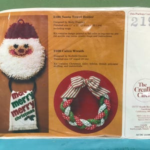May include: A craft kit package featuring a Santa towel holder and a calico wreath. The Santa towel holder has a Santa face design with a towel that says "merry merry christmas". The wreath is red, green, and white.