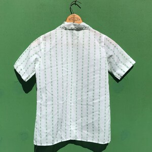 Puede incluir: Una camisa blanca de manga corta con botones y un patr&oacute;n de tr&eacute;bol verde. La camisa tiene cuello y cierre de botones en la parte delantera.