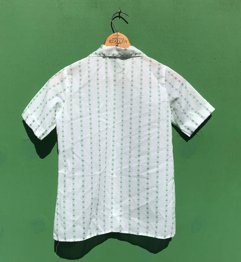 Puede incluir: Una camisa blanca de manga corta con botones y un patr&oacute;n de tr&eacute;bol verde. La camisa tiene cuello y cierre de botones en la parte delantera.