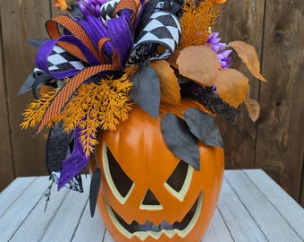 Puede incluir: Una calabaza naranja tallada como centro de mesa, decorada con un arreglo de follaje artificial morado y naranja, hojas negras y un lazo de varias capas con patrones de cuadros blancos y negros y rayas moradas y naranjas.