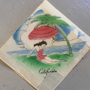 May include: Vintage handkerchief with a beach scene. A woman in a red polka-dot swimsuit reclines under a red umbrella and palm tree. The word "California" is written in cursive. The background features a blue sky and ocean.