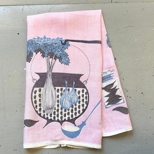 May include: A light pink kitchen towel featuring a dark gray cooking pot with a blue leafy plant inside, two blue onions, a blue ladle, and a glass jar filled with dark gray leaves. The design is printed in a repeating pattern on the fabric.