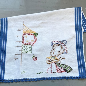 May include: A white tea towel with blue trim and a crocheted edge. The towel features an embroidered design of a person playing a guitar with a red, yellow, and green hat. The design also includes a red, yellow, and green building with a ladder.