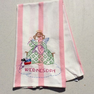 May include: White and pink striped kitchen towel with an embroidered angel design. The angel is wearing a green dress and holding a pink hat. The word "WEDNESDAY" is embroidered below the angel.