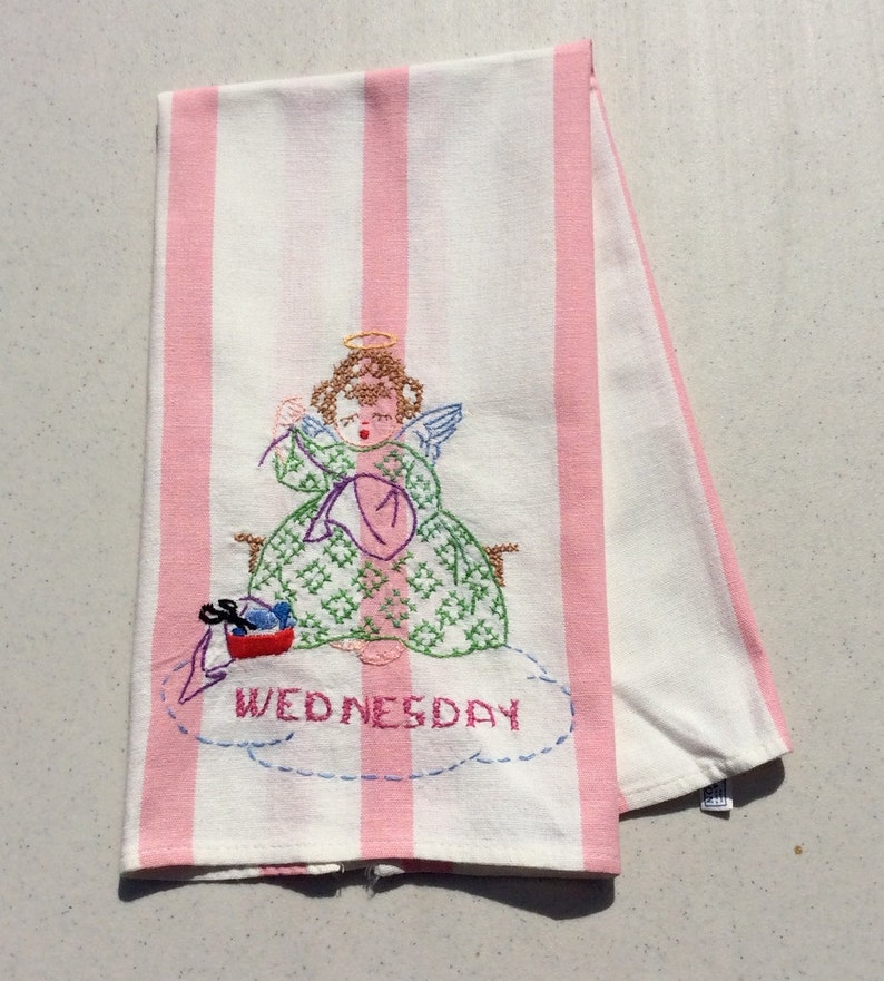 May include: White and pink striped kitchen towel with an embroidered angel design. The angel is wearing a green dress and holding a pink hat. The word "WEDNESDAY" is embroidered below the angel.