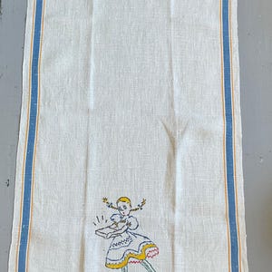 May include: A rectangular white linen kitchen towel featuring vertical blue and yellow stripes along the edges. The center bottom displays an embroidered illustration of a girl with blonde pigtails holding a box, with the word SILVER below.