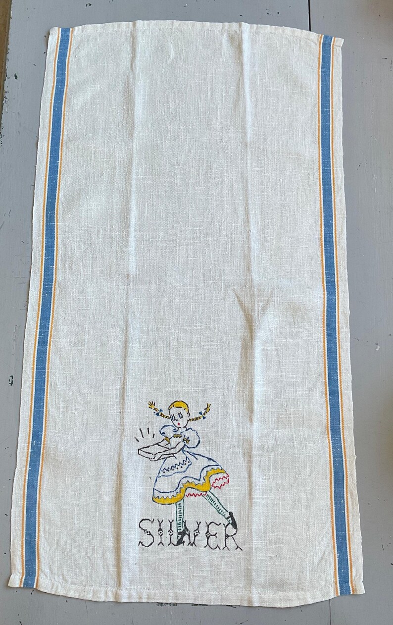 May include: A rectangular white linen kitchen towel featuring vertical blue and yellow stripes along the edges. The center bottom displays an embroidered illustration of a girl with blonde pigtails holding a box, with the word SILVER below.