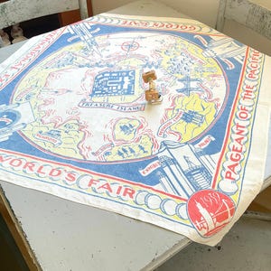 May include: A square souvenir textile featuring a 1939 World's Fair map in blue, red, and yellow. The print includes text reading International Exposition, Pageant of the Pacific, Treasure Island, and 1939, with illustrations of landmarks and bridges.