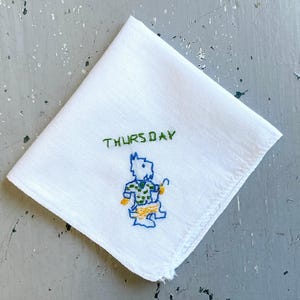 May include: White handkerchief with embroidered design. The word "THURSDAY" is stitched in green above a whimsical figure in blue, green, and yellow. The handkerchief is folded and lies on a gray surface.