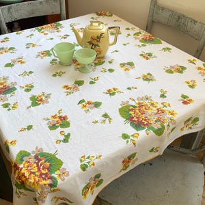 May include: A white tablecloth featuring a repeating floral pattern of yellow, orange, and green pansies. A pale yellow ceramic teapot with a floral design and two light green glass teacups are placed on the centre of the fabric surface.