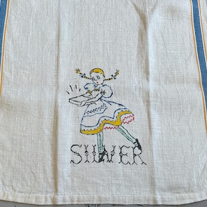 May include: An off-white linen tea towel featuring blue and yellow vertical stripes along the edges. The center displays an embroidered illustration of a girl with blonde pigtails holding a box, with the word SILVER stitched in black below.