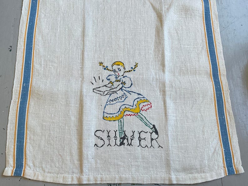 May include: An off-white linen tea towel featuring blue and yellow vertical stripes along the edges. The center displays an embroidered illustration of a girl with blonde pigtails holding a box, with the word SILVER stitched in black below.