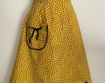 May include: A yellow apron with a repeating pattern of small maroon squares. The apron has a pocket with a black bow and black trim. The bottom edge is scalloped with black trim.
