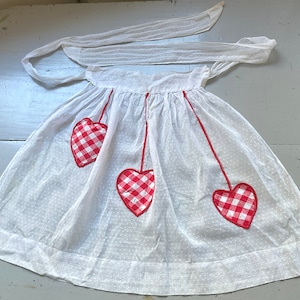 May include: White cotton apron with red and white gingham hearts. The apron has a gathered waist and a tie at the neck.
