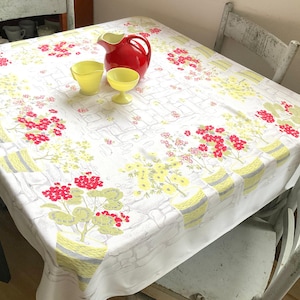 May include: A vintage tablecloth with a floral pattern in red, yellow, and green on a white background. A red pitcher and two yellow glass bowls sit on the table. The tablecloth is on a square table.