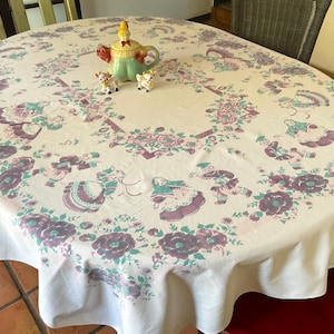 May include: A round white tablecloth featuring a repeating pattern of purple and green floral motifs and figures in traditional clothing. A ceramic teapot and two small animal figurines are placed on the center of the fabric surface.