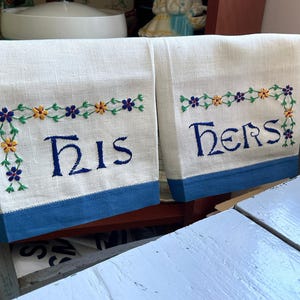 May include: Two white linen tea towels with blue trim and embroidered floral borders. One towel says "His" and the other says "Hers".