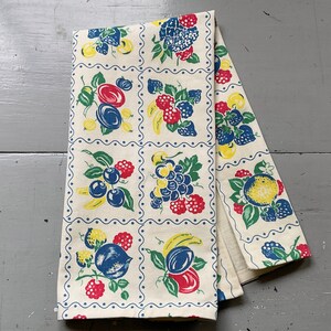 May include: A rectangular kitchen towel featuring a repeating grid pattern of various fruits including red raspberries, blue plums, yellow lemons, and purple grapes. The design is printed on an off-white fabric with blue scalloped borders.