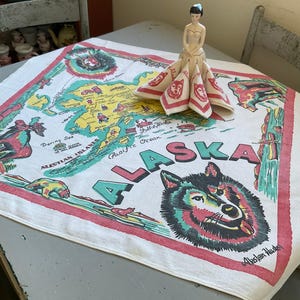 May include: A white cotton tea towel featuring a colourful map of Alaska. The map includes the Bering Sea, Aleutian Islands, Pacific Ocean, and the text "ALASKA". A small porcelain doll wearing a red, white and blue dress is sitting on the towel.