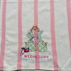 May include: Embroidered hand towel with pink and white stripes, featuring an angel in a green dress and halo, knitting with yarn. The word "WEDNESDAY" is embroidered below the angel on the towel.