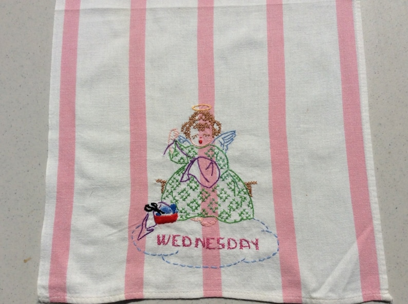 May include: Embroidered hand towel with pink and white stripes, featuring an angel in a green dress and halo, knitting with yarn. The word "WEDNESDAY" is embroidered below the angel on the towel.