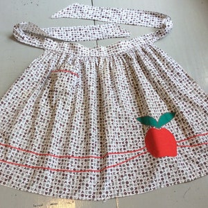 May include: A white apron with a repeating pattern of grey and brown shapes. It features a red apple appliqué with green leaves, a pocket, and red trim. The apron has a matching patterned tie.