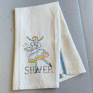 May include: A folded off-white linen tea towel featuring a hand-embroidered illustration of a girl with blonde pigtails wearing a blue and yellow dress, holding a rectangular object, with the word SILVER stitched in black thread below the figure.