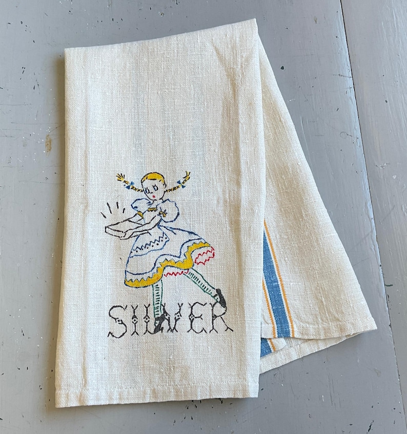 May include: A folded off-white linen tea towel featuring a hand-embroidered illustration of a girl with blonde pigtails wearing a blue and yellow dress, holding a rectangular object, with the word SILVER stitched in black thread below the figure.