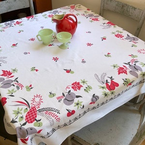May include: A white tablecloth featuring a vibrant fruit and floral design with red pears, grey plums, and green apples. A red jug and two green cups are on the table. The tablecloth covers a square table with two grey chairs.