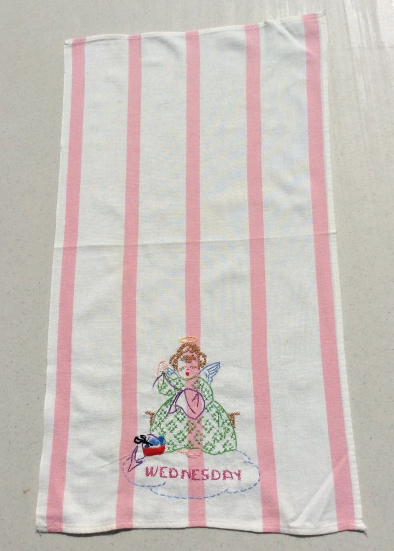 May include: White and pink striped tea towel featuring an embroidered angel design. The angel is wearing a green dress and holding a pink heart. The word "WEDNESDAY" is embroidered below the angel.