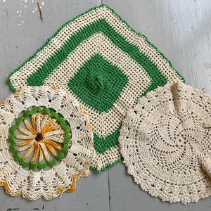 Peut inclure: Trois napperons en crochet faits à la main : une pièce carrée avec des rayures alternées vertes et crème, une pièce circulaire avec un centre de fleur jaune et orange, et une pièce circulaire crème avec un motif en spirale ajouré.