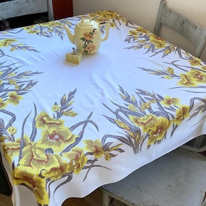 May include: A white tablecloth with a yellow floral pattern covers a table. A yellow teapot with floral accents and a small decorative item sit on the tablecloth. The table is surrounded by two distressed white chairs.