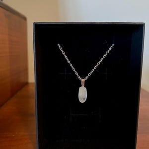 May include: A silver chain necklace with a small, white, oval-shaped pendant. The necklace is displayed in a black velvet box.