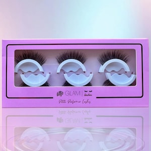 May include: A pink box containing three sets of false eyelashes. The box has a black border and the text "little GLAM lashes Petite Performer Lashes". Each set of lashes is held in place by a white, curved holder.