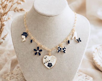 May include: Gold-tone necklace with multiple charms, including a hat, flower, bow, and high heel. The central charm is a white heart with a black floral design and the number 5. The necklace is displayed on a mannequin.
