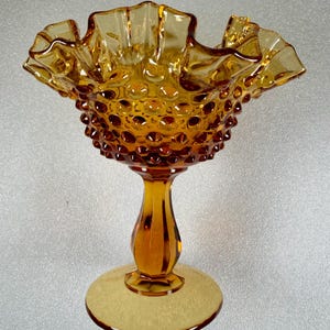May include: An amber-colored glass compote dish featuring a hobnail texture on the bowl and a ruffled, crimped rim. The piece stands on a smooth, tapered pedestal base that connects to a circular foot, showcasing a translucent, warm orange hue.