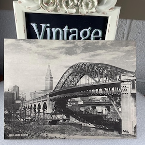 May include: A black and white vintage postcard featuring a bridge with a large arch and a city skyline. The postcard is displayed in a white frame with floral embellishments and the word "Vintage". The text "HIGH LEVEL BRIDGE" is visible.