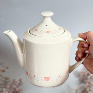 Könnte beinhalten: Weiße Teekanne aus Keramik mit einem rosa-grünen Blumendesign. Die Teekanne hat einen Deckel mit einem kleinen Loch oben.