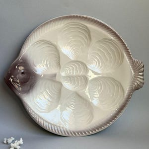 May include: A white ceramic oyster plate shaped like a fish with eight individual oyster shells. The plate has a purple rim and a purple fish head.