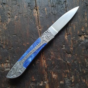 Puede incluir: Un cuchillo artesanal con una hoja de acero pulido y un mango azul y plateado. El mango presenta intrincados grabados florales. El cuchillo se muestra sobre una superficie de madera oscura y texturizada.