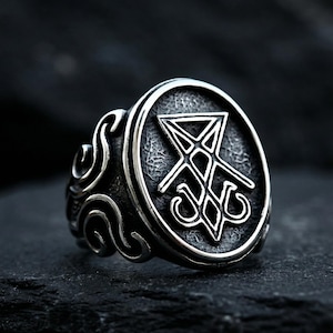 Könnte beinhalten: Silberfarbener Ring mit ovaler Oberfläche, die ein geometrisches Symbol zeigt. Der Ring hat eine strukturierte Oberfläche und verzierte Schnörkel an den Seiten. Der Hintergrund ist dunkel.