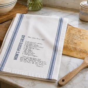 May include: A white kitchen towel featuring two vertical blue stripes and the printed text "MOM'S POTATO SALAD" followed by a handwritten-style recipe for potato salad. The towel is folded and placed on a wooden surface next to a wooden spoon.