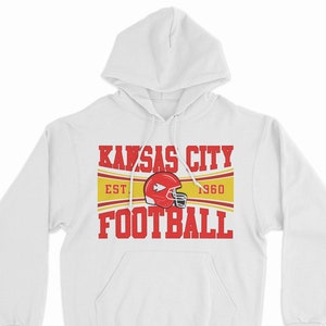 May include: White hooded sweatshirt with a red and yellow graphic that says "Kansas City Football" with the team logo and "Est. 1960" underneath.