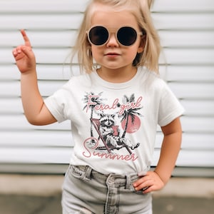 May include: A white t-shirt with a graphic of a raccoon sitting in a beach chair with a drink. The text "Feral girl summer" is printed in pink on the shirt.