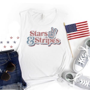 May include: White tank top with a vintage-style graphic that says "Stars & Stripes" in red, white and blue. The graphic includes a peace sign with red and white stripes. The tank top is laid out on a white surface with a pair of grey sneakers, a pair of denim shorts and a small American flag.