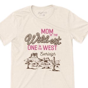May include: A cream-coloured t-shirt featuring the text "MOM of the Wildest ONE in the WEST Everleigh" in a western-style font. The design includes a desert landscape with cacti and mountains.